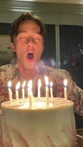 A man celebrating his birthday by blowing out candles on a cake, with cheerful birthday memes in the background.